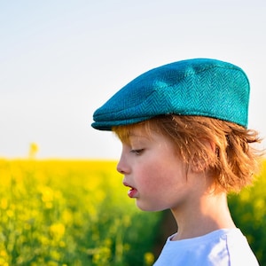 May include: A teal herringbone flat cap on a person with red hair and a white shirt. The background is a field of yellow flowers under a clear sky.