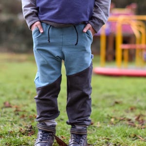 May include: A child's casual pants with a two-tone design. The upper portion is a light blue, while the lower legs are dark gray. The pants have a drawstring waist and are paired with dark blue boots. The background is a grassy area.