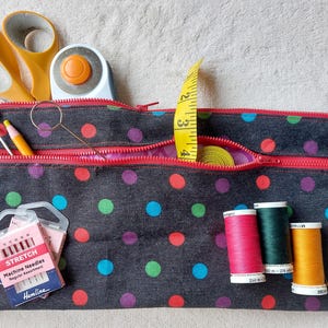 May include: A dark grey fabric pouch with a red zip and colourful polka dots. It contains a yellow measuring tape, orange scissors, a rotary cutter, a seam ripper, three spools of thread in pink, green, and yellow, and a pack of Hemline stretch machine needles.