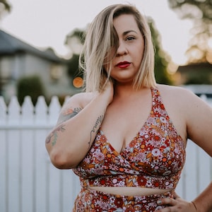 Puede incluir: Una mujer con un top halter con estampado floral y pantalones de cintura alta a juego. El conjunto presenta un fondo rojo con un patrón de pequeñas flores en tonos rosa, naranja y blanco. Está de pie cerca de una valla blanca.