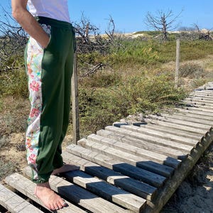 Könnte beinhalten: Grüne Jogginghose mit floralem Seitenpanel. Das Blumenmuster zeigt rosa und orangefarbene Blumen auf weißem Grund. Die Hose hat einen Kordelzug in der Taille und Taschen. Die Person steht auf einem Holzsteg.