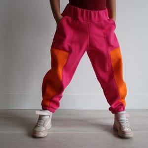 May include: Bright pink and orange colour-block trousers with pockets. The trousers have an elastic waistband and are cuffed at the ankles. The trousers are paired with white and pink trainers. The person is standing with their hands in the pockets.