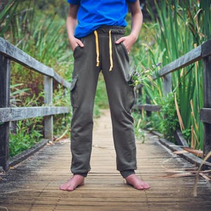 May include: A person wearing olive green cargo sweatpants with yellow drawstrings and a blue shirt stands barefoot on a wooden bridge. The pants have pockets on the sides. The background features a wooden fence and tall green plants.