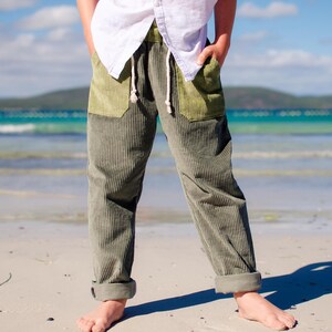May include: Olive green corduroy pants with a drawstring waist and olive green pockets. The pants are cuffed at the ankles. The person is standing on a sandy beach with the ocean in the background.