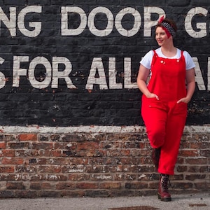 Puede incluir: Una persona con un mono rojo y una camiseta blanca posa frente a una pared de ladrillos con texto pintado. La persona también lleva una diadema de lunares y botas marrones. La pared muestra las palabras "RIDING DOOR GEAR" y "TOOLS FOR ALL TRADES".