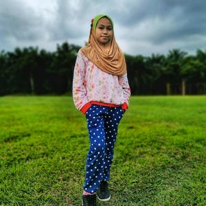 Puede incluir: Una persona está de pie en un campo cubierto de hierba, vistiendo una parte superior de manga larga de color rosa claro y blanco con un patrón y ribete rojo, combinada con pantalones azules cubiertos de estrellas blancas. También llevan zapatillas negras y un hiyab marrón con un tocado verde.