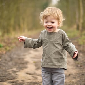 May include: A young child with blond hair laughs while walking on a dirt path in a forest. The child wears a long-sleeved olive green shirt and gray pants. The background is blurred with trees.