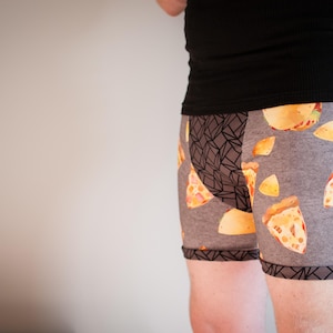 May include: Gray boxer briefs with a pizza slice pattern. The briefs have a geometric design on the side and a black trim. The person is wearing a black tank top.