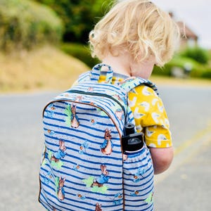 May include: A blue and white striped backpack featuring a Peter Rabbit design. The backpack has a side pocket holding a black water bottle. The child is wearing a yellow shirt with a bird pattern.