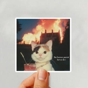May include: A square sticker featuring a cat wearing a tiara and necklace, holding a drink with a straw. The background shows a burning building. Text on the sticker reads "the horrors persist but so do I."