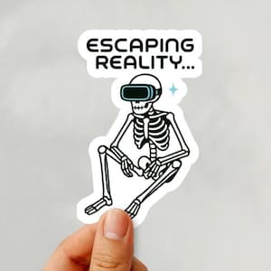 May include: A white sticker featuring a black and white illustration of a skeleton wearing VR goggles. The text "ESCAPING REALITY..." is above the skeleton. A hand holds the sticker against a white background.
