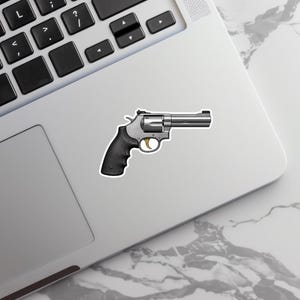 May include: A sticker featuring a silver revolver with a black grip and a gold trigger. The sticker is placed on a laptop, with the keyboard visible in the background. The revolver is a classic firearm design.