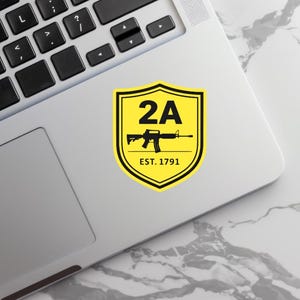 May include: A yellow and black sticker with a shield design. The sticker features the text "2A" above a black rifle graphic and the text "EST. 1791". The sticker is placed on a laptop.
