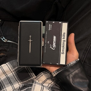 May include: A black leather cigarette case with a silver sword charm and a black cigarette box. The cigarette box has the text "Smoking kills" and "Cigarronne Imperial Collection". The background is a plaid shirt and black denim.