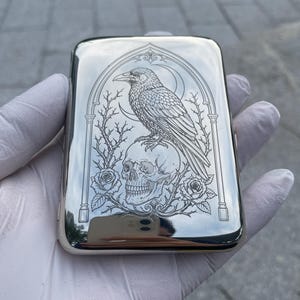 Op de afbeelding: Een rechthoekige, zilverkleurige metalen houder met een gedetailleerd lasergegraveerd ontwerp van een raaf die op een menselijke schedel zit. Het kunstwerk is omlijst door een gotische boog, geflankeerd door doornige rozenranken en een maansikkel.