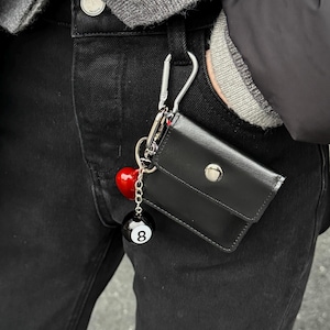 May include: A black leather wallet with a silver snap closure is attached to a belt loop with a silver carabiner clip. A red heart and a black 8-ball charm dangle from the wallet.