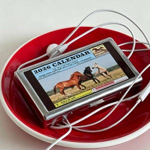 Könnte beinhalten: Ein silbergerahmter Kalender für 2026 mit drei Pferden auf einer Wiese, der in einer roten Schüssel liegt. Weiße kabelgebundene Ohrhörer sind um den Kalender drapiert. Der Kalendertext lautet "Holen Sie sich Ihr Glück mit diesem Glückskalender!"