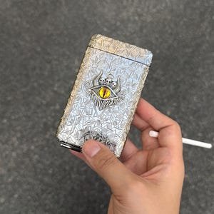 May include: Silver cigarette case with an ornate design. The case features a textured surface and a central emblem of an eye with a crown. A lit cigarette is held nearby.