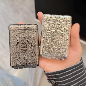 Puede incluir: Dos estuches rectangulares de metal con tapa abatible, con intrincados diseños en relieve de flores y figuras aladas. El centro de cada estuche muestra un medallón ovalado con escritura árabe grabada. Uno tiene un acabado oscuro y el otro plateado.