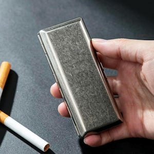 May include: A rectangular, metallic cigarette case with a textured surface is held in a hand. The case is silver-toned and has a hinged lid. Two cigarettes are visible to the left of the case, one lit and one unlit.