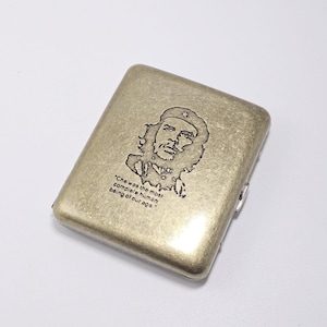 Könnte beinhalten: Eine rechteckige, metallisch goldene Zigarettenetui. Das Etui zeigt ein schwarz graviertes Porträt von Che Guevara und das Zitat: „Che war der vollkommenste Mensch unserer Zeit.“