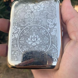 Könnte beinhalten: Ein rechteckiges Zigarettenetui aus silberfarbenem Metall mit einer detaillierten Gravur. Die Oberfläche zeigt ein zentrales kreisförmiges Medaillon mit einem Blumenmotiv, umgeben von geometrischen Mustern und floralen Ranken.