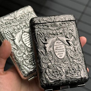 May include: Two ornate metal cigarette cases. One is silver, the other is dark grey. Both feature detailed floral and angel designs with text. The cases have a rectangular shape with a hinged lid and a small metal clasp.