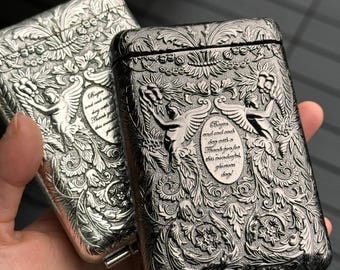 May include: Two ornate metal cigarette cases. One is silver, the other is dark gray. Both feature detailed floral and angel designs with text. The cases have a rectangular shape with a hinged lid and a small metal clasp.