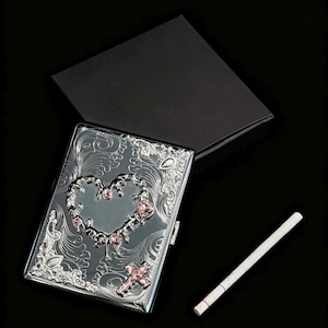 K&ouml;nnte beinhalten: Silberne Zigarettenetui mit herzf&ouml;rmigem Design, verziert mit rosa Edelsteinen und floralen Mustern. Das Etui ist ge&ouml;ffnet und zeigt eine Zigarette. Eine schwarze Box befindet sich hinter dem Etui.