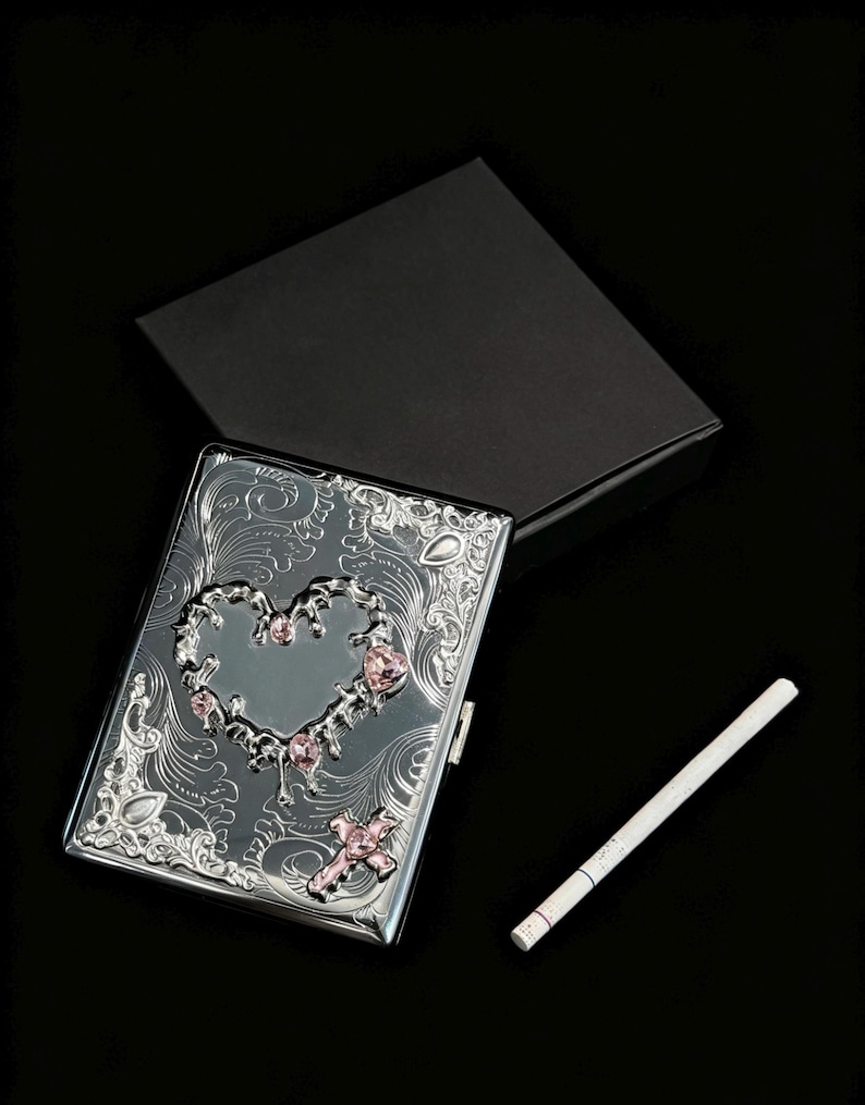 K&ouml;nnte beinhalten: Silberne Zigarettenetui mit herzf&ouml;rmigem Design, verziert mit rosa Edelsteinen und floralen Mustern. Das Etui ist ge&ouml;ffnet und zeigt eine Zigarette. Eine schwarze Box befindet sich hinter dem Etui.