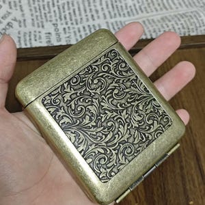 Könnte beinhalten: Eine Zigarettenetui aus Messing im Vintage-Stil mit einem verzierten floralen Muster auf dem Deckel. Die Hülle hat eine rechteckige Form mit abgerundeten Ecken und einer Scharnieröffnung. Die Hülle wird in einer Hand gehalten.