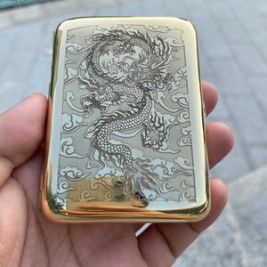 Op de afbeelding: Een rechthoekig sigarettendoosje van goudkleurig metaal met een gedetailleerde lasergravure van een draak omringd door gestileerde wolken. Het doosje heeft een gepolijste, reflecterende rand en een mat middenpaneel met de drakenafbeelding.