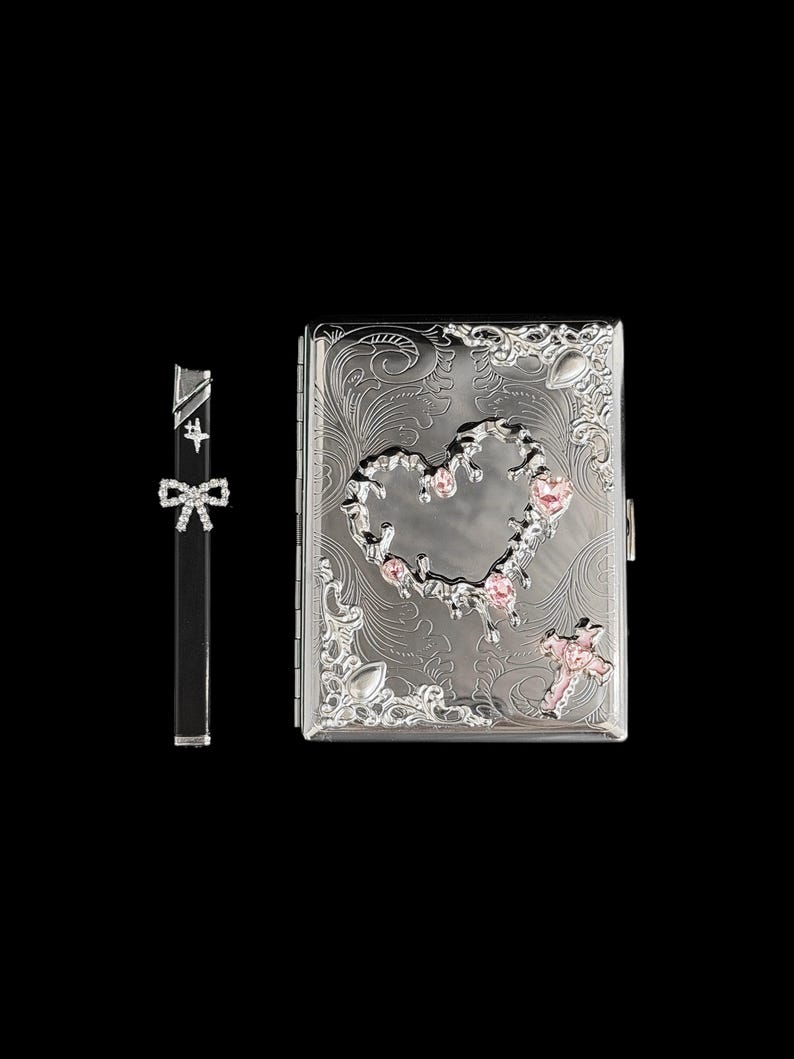K&ouml;nnte beinhalten: Eine silberne Zigarettenetui und ein passendes Feuerzeug. Die Etui hat ein eingraviertes Blumenmuster mit herzf&ouml;rmigem Ausschnitt und rosa Edelsteinakzenten. Das Feuerzeug ist schwarz mit einer Schleife.