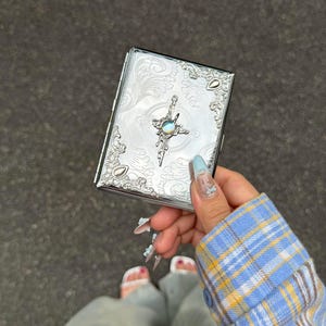 Peut inclure: Un étui rectangulaire en métal argenté présentant un motif complexe de volutes en relief sur toute la surface. Le centre est orné d'un emblème en forme de croix contenant une petite pierre circulaire irisée sertie au milieu.