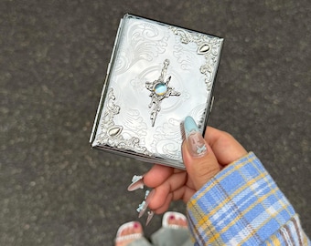May include: A rectangular silver-toned metal case featuring an intricate embossed scrollwork pattern across the surface. The center is adorned with a cross-shaped emblem containing a small, iridescent, circular stone set in the middle.