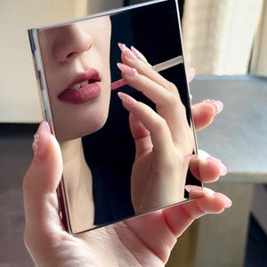 Puede incluir: Un espejo compacto cuadrado plateado que refleja a una persona aplicándose lápiz labial. Los labios de la persona son de un rojo intenso, y su mano, con uñas largas, rosas y decoradas, sostiene el espejo. El marco del espejo es plateado brillante.