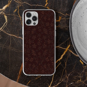May include: A maroon phone case with a floral pattern. The case has a clear border and a camera cutout. The floral design is in a lighter shade of maroon, creating a subtle contrast. The phone case is on a dark marble surface.