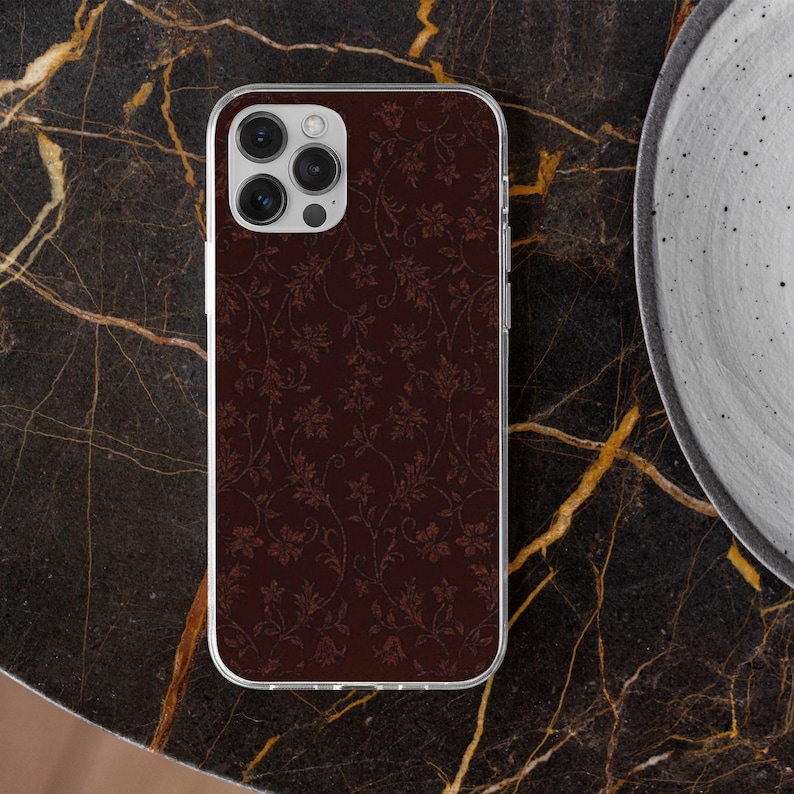 May include: A maroon phone case with a floral pattern. The case has a clear border and a camera cutout. The floral design is in a lighter shade of maroon, creating a subtle contrast. The phone case is on a dark marble surface.
