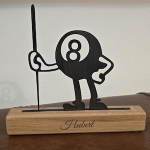Puede incluir: Figura de madera negra de una bola de billar con el número 8, sosteniendo un taco. Montada sobre una base de madera marrón claro con el nombre "Hubert" grabado. Un objeto decorativo.