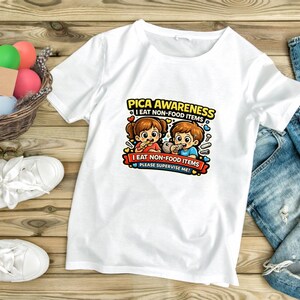Könnte beinhalten: Weißes T-Shirt mit einem farbenfrohen Aufdruck. Der Aufdruck lautet "PICA AWARENESS I EAT NON-FOOD ITEMS PLEASE SUPERVISE ME!" und zeigt Cartoon-Kinder. Das Shirt liegt auf einer Holzoberfläche.