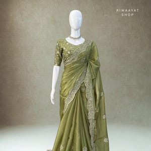 May include: An olive green saree with intricate silver embroidery on the borders and bodice. The saree is draped on a mannequin, showcasing its flowing fabric and design. The text "RIWAAYAT SHOP" is visible.