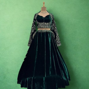 May include: A dark green velvet outfit, comprising a crop top, long-sleeved jacket, and a flowing skirt. The jacket and top have detailed silver embroidery. The ensemble is displayed on a mannequin against a green background. The text "RIWAAYAT SHOP" is visible.
