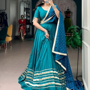 May include: A teal lehenga choli set featuring a pleated floor-length skirt with horizontal gold metallic bands at the hem. The matching blouse has short sleeves with tassel details, and the teal dupatta is adorned with small white dots and gold trim.