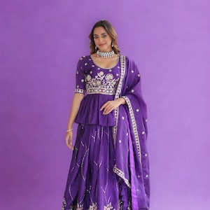Puede incluir: Un conjunto morado bordado con un dupatta a juego. La parte superior tiene un diseño peplum con intrincados bordados florales y una cintura con cuentas. La falda también presenta bordados detallados. El conjunto se usa con un collar plateado.