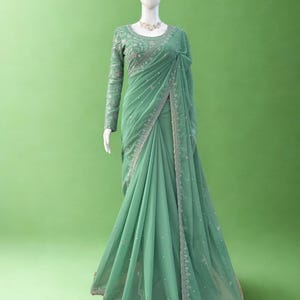Puede incluir: Un sari verde salvia con intrincados bordados y abalorios, exhibido en un maniquí. La parte superior de manga larga presenta patrones florales detallados, y el borde del sari está adornado con detalles plateados. El texto "RIWAAYAT SHOP" es visible.
