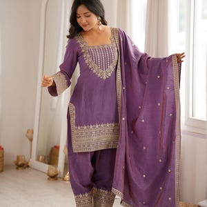 Può includere: Un completo salwar kameez viola con scollo quadrato, maniche lunghe e intricati ricami dorati sul corpetto, sui polsini e sull'orlo. I pantaloni e il dupatta abbinati sono decorati con motivi dorati. Il testo recita RIWAAYAT SHOP.