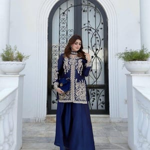 May include: A navy blue long-sleeved tunic and wide-leg trousers set featuring intricate cream-colored embroidery along the front panels and cuffs. The tunic has a high neckline and is paired with a matching navy blue handbag with a gold logo.