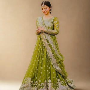 Peut inclure: Une longue robe vert olive avec des broderies et des ornements argentés complexes. La robe a des manches longues et un châle assorti. L'image comprend le texte "RIWAAYAT SHOP".