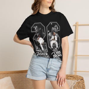 May include: A black short-sleeve t-shirt featuring a large graphic print of Usher Raymond and Chris Brown. The design includes the number 23, black and white portraits, and full-body images of both artists in white suits with their names printed below.