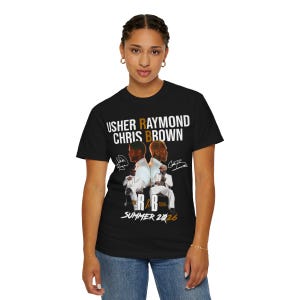 May include: A black short-sleeve t-shirt featuring a graphic print of Usher Raymond and Chris Brown. The text reads USHER RAYMOND CHRIS BROWN, THE R&B TOUR, and SUMMER 2026. The design includes two figures in white suits and two signatures on a black background.